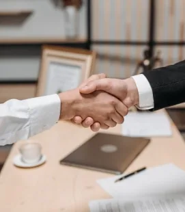 Two professionals in business attire shaking hands across a desk in an office setting
